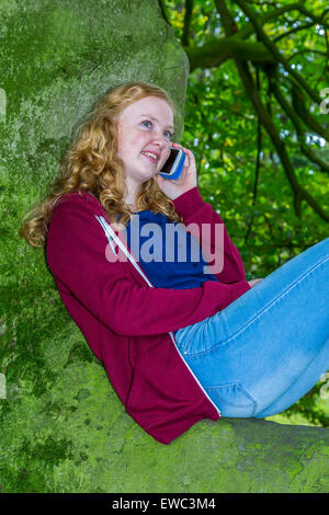 Teenage girl sitting on tree calling and smiling with mobile phone Stock Photo