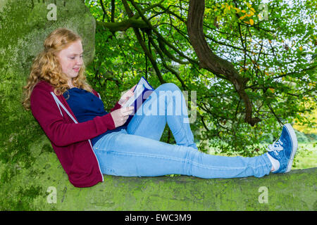 Caucasian teenage girl with red hair lying reading book in green tree Stock Photo