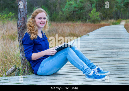 European teenage girl reading book on wooden path in forest Stock Photo