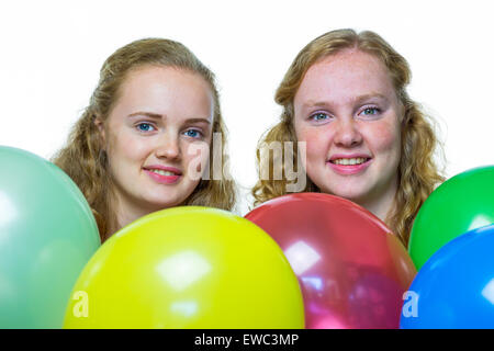 Two smiling caucasian teenage girls behind various colored balloons isolated on white background
