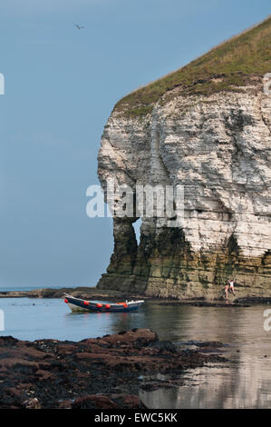 Scenic view in sunny summer, of blue sky, calm sea & wooden boat moored off-shore by arch in towering chalk cliffs. Flamborough, Yorkshire coast, UK. Stock Photo