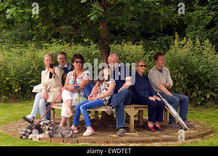 Group of people (& 2 dogs) sitting on a bench under a tree watching a local summer community parade, Frensham, Surrey, UK. Stock Photo