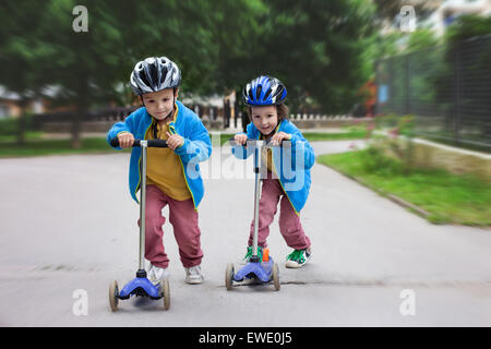 Two cute boys, compete in riding scooters, outdoor in the park, summertime Stock Photo