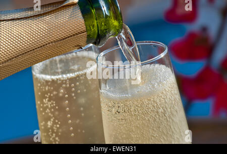 POURING SPARKLING WINE ALFRESCO Close view on pouring chilled glasses of champagne on sunlit terrace with Bougainvillea flowers and pool in background Stock Photo