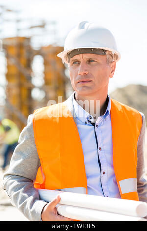Thoughtful architect looking away while holding blueprints at construction site Stock Photo