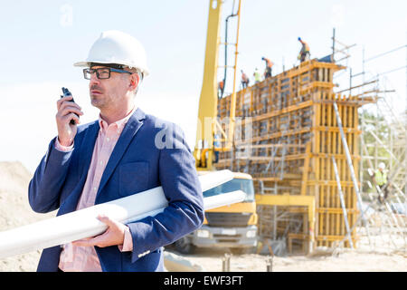 Architect using walkie-talkie while holding blueprints at construction site Stock Photo