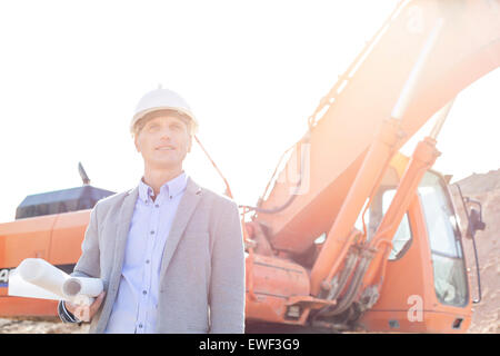 Thoughtful engineer looking away while holding blueprints by bulldozer at construction site Stock Photo