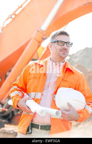 Thoughtful architect holding hardhat and blueprints at construction site Stock Photo