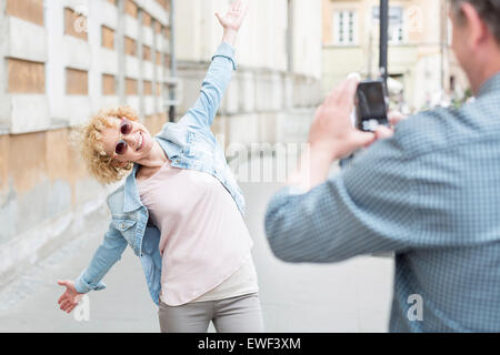 Man photographing playful woman standing with arms outstretched on city street Stock Photo