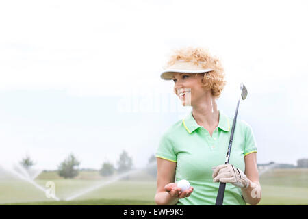 Happy middle-aged woman looking away while holding golf club and ball Stock Photo