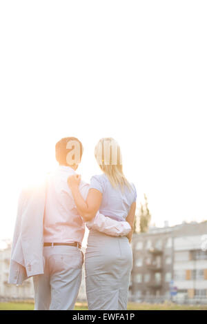 Rear view of business couple standing with arms around against clear sky on sunny day Stock Photo