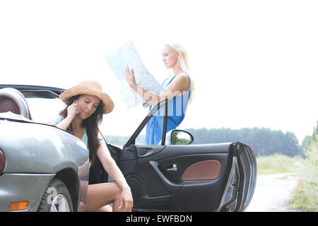 Woman using cell phone in convertible while friend reading map on road Stock Photo