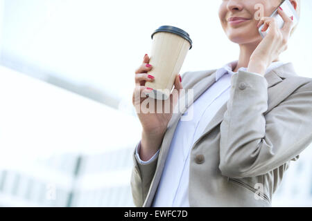 Midsection of businesswoman using cell phone while holding disposable cup outdoors Stock Photo