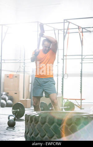 Determined man hitting tire with sledgehammer in crossfit gym Stock Photo