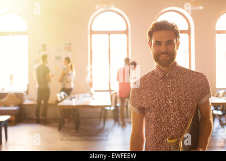 Portrait smiling casual businessman with skateboard in sunny open office Stock Photo