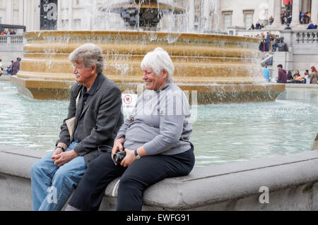 Happy senior couple sitting on the fountain at Trafalgar Square, London England United Kingdom UK Stock Photo