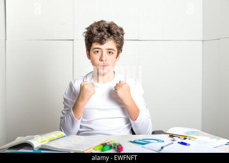 Caucasian smooth-skinned boy sits in front of homework: he is calm and confident and makes a gesture of success raising fists on industrial background Stock Photo