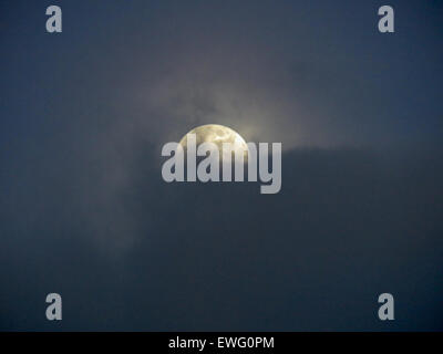 A full moon peeks through a blanket of clouds, partially obscured. The soft glow of the moon illuminates the clouds, creating an ethereal scene in the night sky. Stock Photo