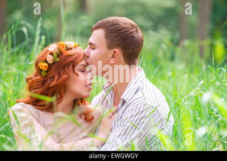 Portrait of beautiful gentle loving couple sitting on the grass in forest, romantic guy with pleasure kissing his girlfriend Stock Photo