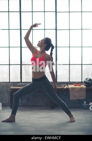 Looking up at her hand, an athletic, strong woman stretches out in a yoga pose in a city loft gym. Stock Photo