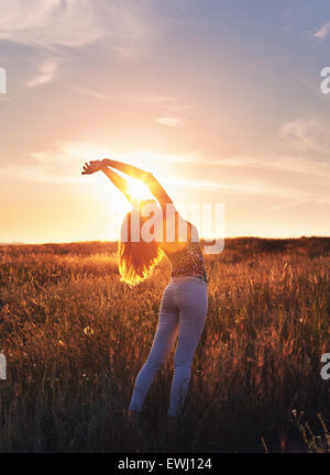 Free happy young woman raising arms at sunset in the field in summer. Background Stock Photo