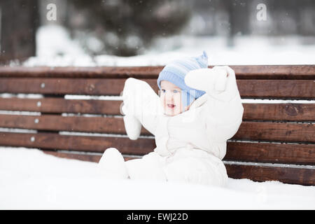 Cute funny baby sitting on a bench in a park on a snowy winter day Stock Photo