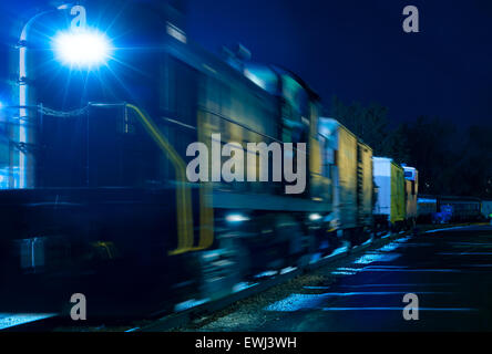 An old diesel locomotive and freight cars glow in an eerie blue under the night lights of a railyard Stock Photo