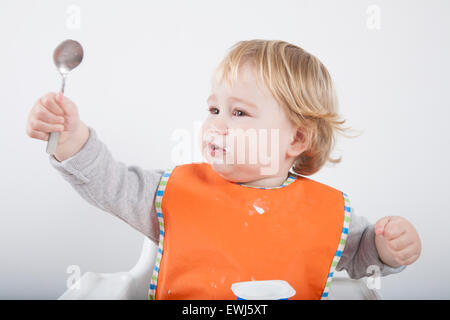 blonde caucasian baby seventeen month age orange bib grey sweater on white high chair with spoon hand up Stock Photo