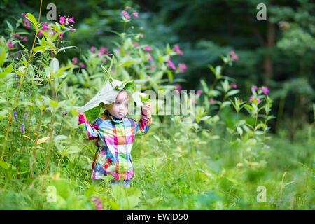 Funny baby girl playing peek a boo in a park under huge leaves