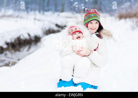 Cute boy hugging his baby sister in a beautiful winter park under a Christmas tree in snow both wearing warm white jackets Stock Photo