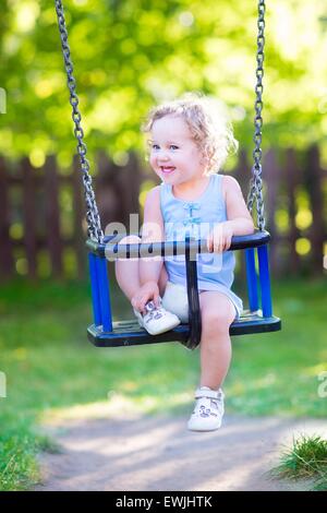 Beautiful baby girl with curly hair wearing a blue dress having fun on a swing on a playground on a sunny summer day Stock Photo