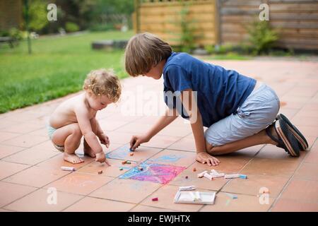 Brother and his baby sister playing together in the backyard painting with colorful chalk Stock Photo