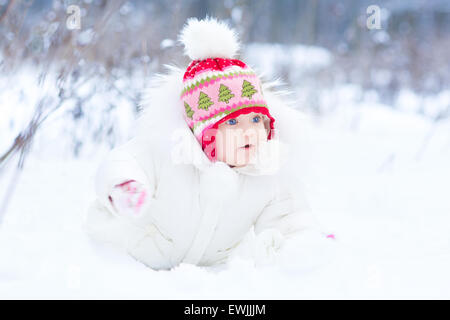 Cute funny little baby wearing a warm white winter jacket and a red hat sitting under a christmas tree in snow Stock Photo