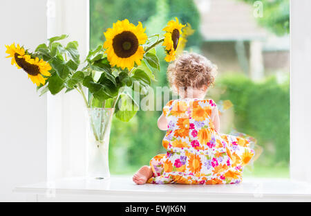 Cute curly baby girl watching out of a window into the garden sitting next to a sunflower bouquet Stock Photo