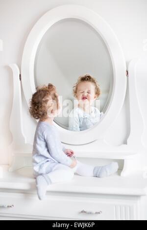 Very funny baby girl with curly hair looking at her reflection in a beautiful white bedroom with a classic dresser with a round Stock Photo