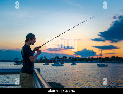 Port Washington, New York, USA. 26th June 2015.  A young man, wearing cap backwards, is fishing as sunset approaches, on the Town Dock on the shores of Manhasset Bay in the North Shore village on Long Island Gold Coast. Credit:  Ann E Parry/Alamy Live News Stock Photo