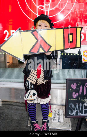 Japan, Osaka. Young Japanese woman dressed in black punk/Gothic style with black hat holding large sign outside store, pointing to way in. Stock Photo