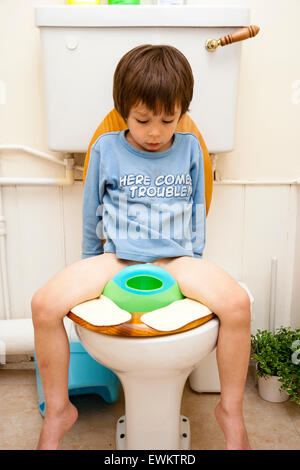 Caucasian young child, boy, 4-5 year old, facing viewer, sitting on baby seat on top of toilet during toilet training. Looking down between his legs. Stock Photo