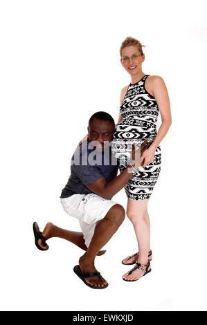 An African American man kneeling before his Caucasian wife, listens on her baby belly, isolated for white background. Stock Photo