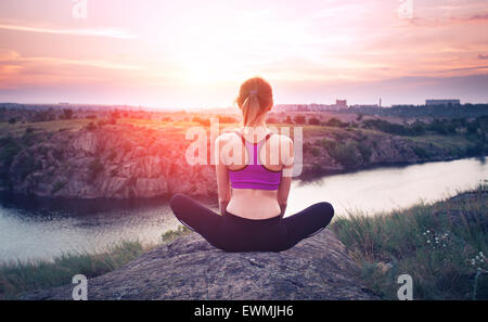 Young woman practicing yoga on the hill at sunset near the river. Stock Photo