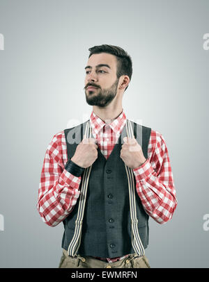 Desaturated portrait of standing young hipster model looking away with raised eyebrow while holding his pants suspenders. Stock Photo