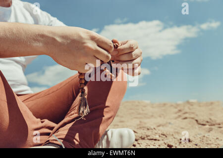 Young hipster man holds a rosary in his hands, sitting on the ground in a sunny summer day Stock Photo