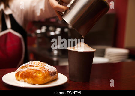 Barista, waitress in red apron making in Takeaway cup Coffee with foam and Cinnamon and sweet Bun on plate Stock Photo