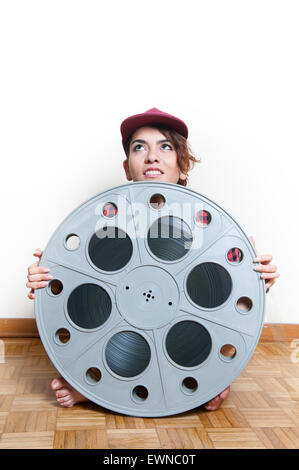 Young smiling woman with red cap sits on wooden floor behind big original cinema movie reel Stock Photo