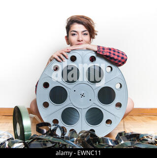 Young smiling woman sits on wooden floor behind big original cinema movie reel and filmstrip Stock Photo