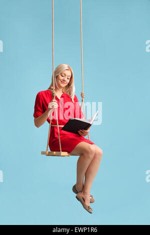 Vertical shot of a young blond woman in a red dress reading a book seated on a swing on blue background Stock Photo