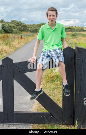 Caucasian teenage boy sitting on black wooden fence in nature as tourist enjoying vacation Stock Photo