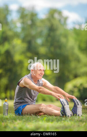 Vertical shot of a senior man stretching his legs seated by a pond in a park and listening to music on headphones Stock Photo