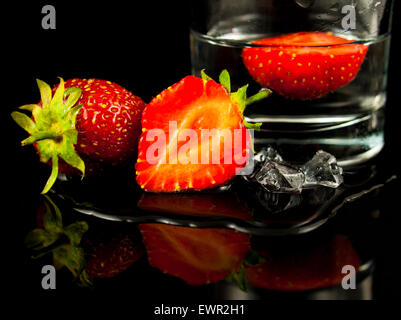 Strawberries, whole, sliced, in glass, water and ice on black reflective background Stock Photo