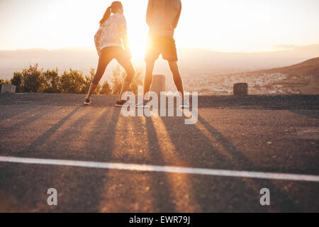 Rear view of young couple standing on hillside in morning facing the sun light. Young woman stretching her legs during a running Stock Photo